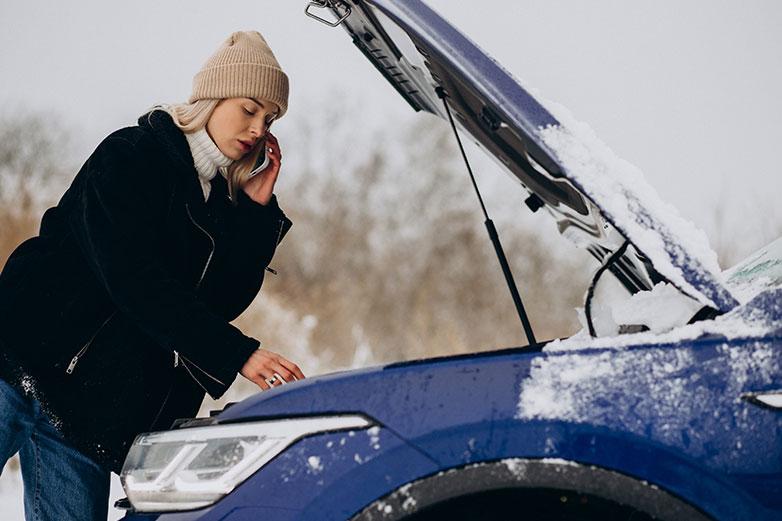 Mujer al tel&eacute;fono, revisando la bater&iacute;a del coche en invierno